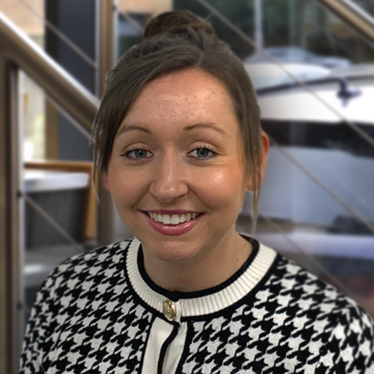 A person wearing a black and white patterned jacket standing indoors near a metal and wooden staircase with glass windows and parked cars visible outside.