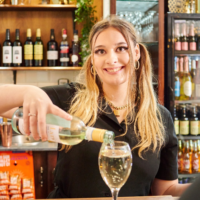 Pub staff member pouring a glass of white wine behind the bar, illustrating Staff, Slips and Safety Risks for Pub Tenants.
