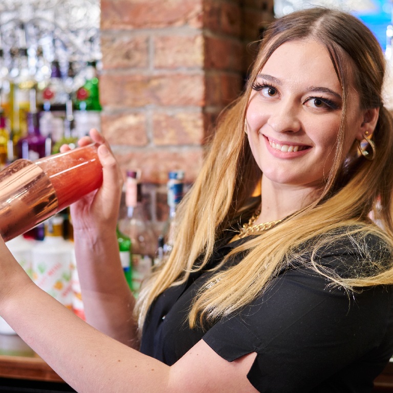 Bartender smiling while shaking a cocktail behind a busy bar in a pub, for a Pub insurance FAQs page.
