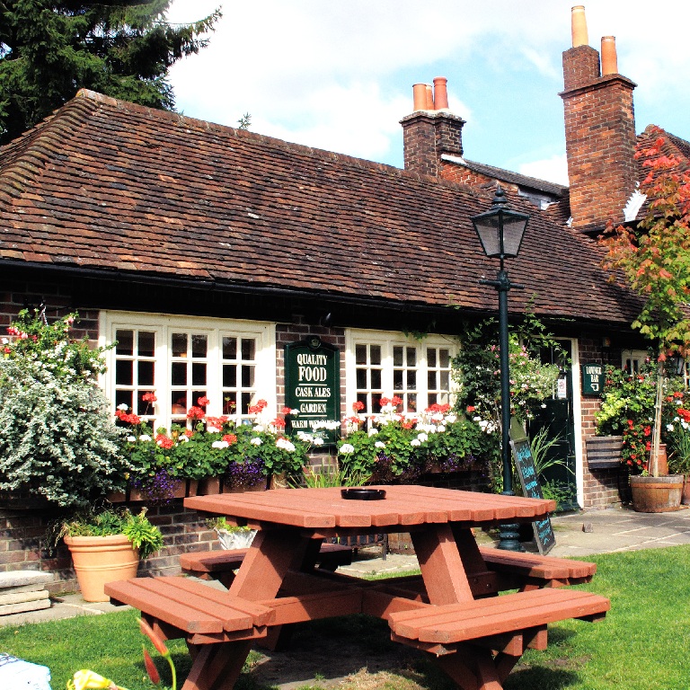 Traditional brick pub with flower boxes, chimney stacks, and a wooden picnic table in a sunny beer garden, for beer garden and pub events insurance cover content.