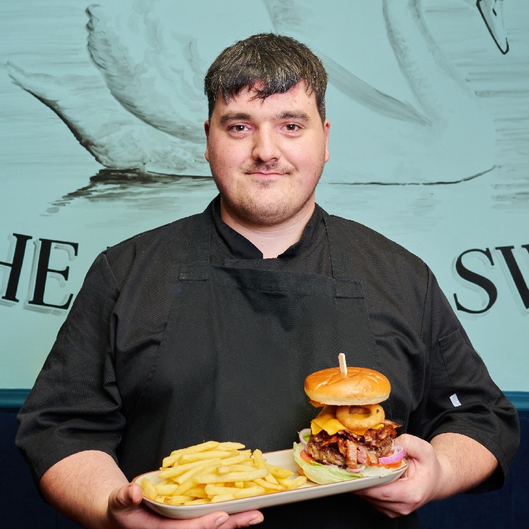 Chef standing indoors at The Swan pub holding a burger, illustrating how much does pub insurance cost.