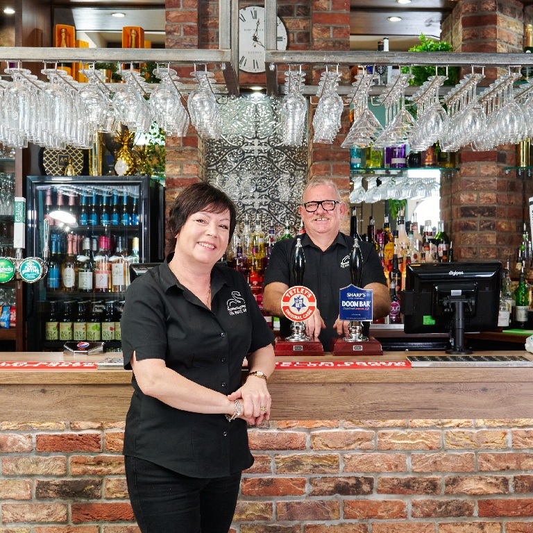 Pub insurance renewal scene with a woman standing at a pub bar and a bartender behind her, with bottles and hanging glassware visible.