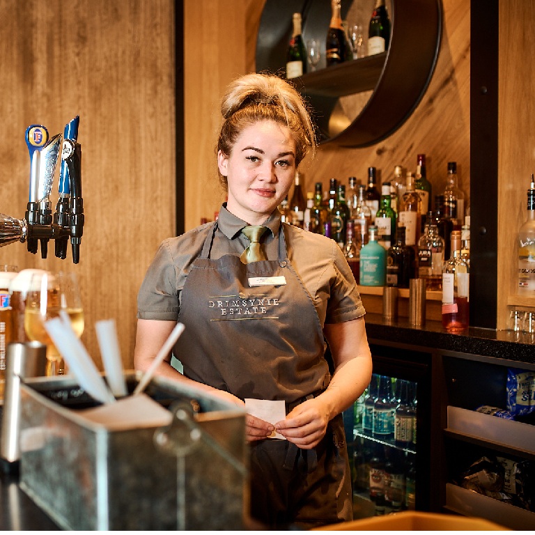 Leasehold vs freehold pub concept: bartender standing behind a modern wooden pub bar with beer taps, glasses, and bottles on display.