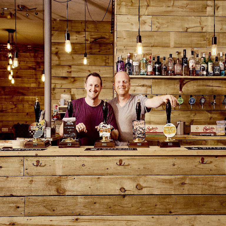 Two men standing behind a wooden bar in a rustic pub with beer pumps, bottles, and warm hanging lights, used to illustrate pub security.