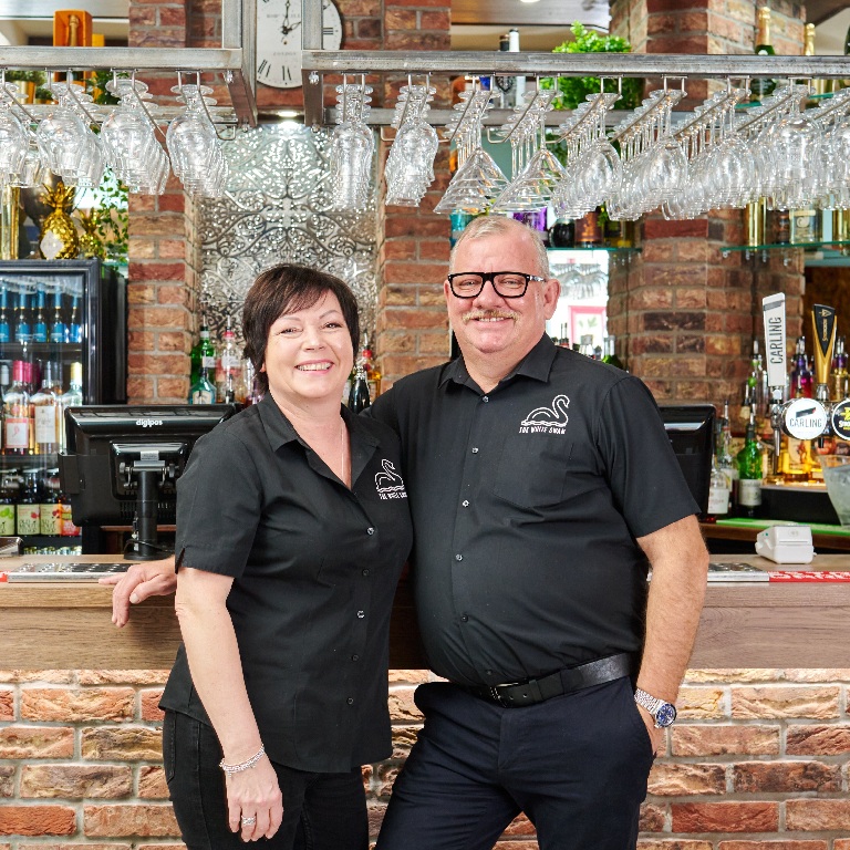 Two pub staff members standing at a bar with beer taps and hanging glasses, illustrating a practical insurance review checklist for landlords, pubcos and tenant operators.