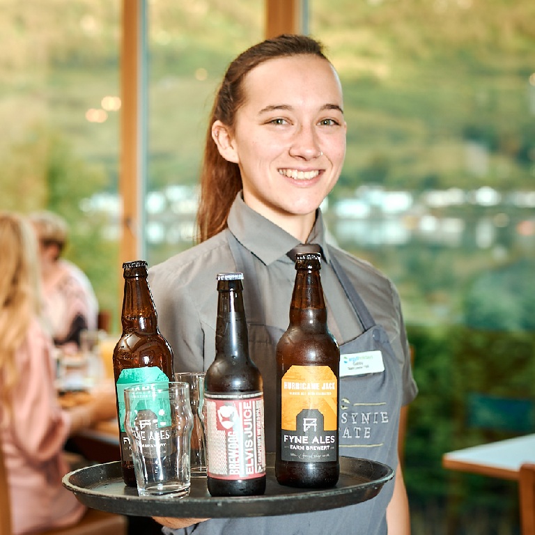 Server carrying bottled drinks on a tray in a restaurant dining area, illustrating restaurant health and safety in a clean, professional setting.