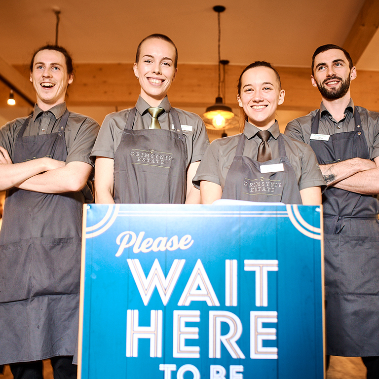 Hospitality risk management team members standing in a restaurant waiting area, with a “Please Wait Here” sign in the foreground.