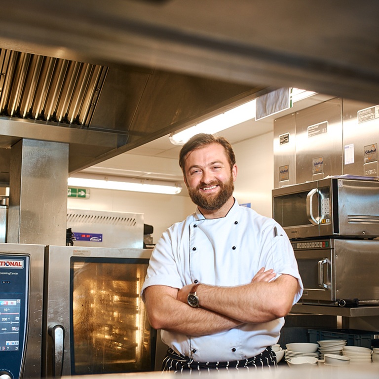 Chef standing with arms crossed in a restaurant kitchen, with stainless steel ovens, shelves, and ventilation equipment around him.