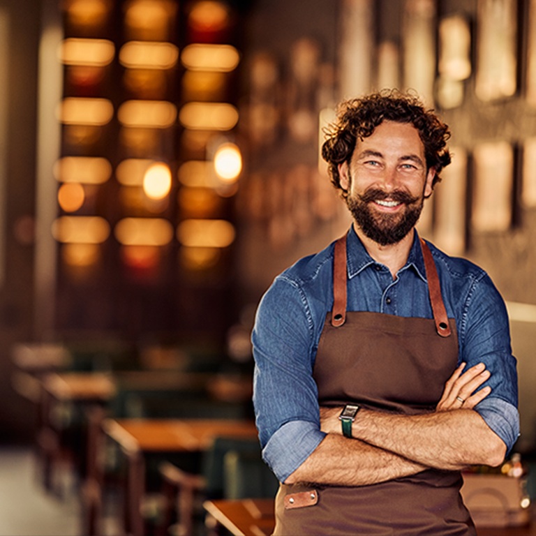 Smiling independent restaurant owner wearing an apron and standing with arms crossed inside a restaurant, illustrating Restaurant Insurance: What Independent Owners Need to Know