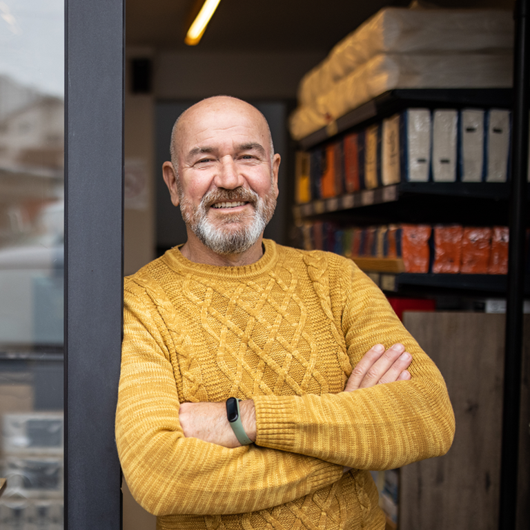 A small business owner wearing a yellow sweater stands confidently with arms crossed in the doorway of a shop displaying cleaning and personal care products on wooden shelves
