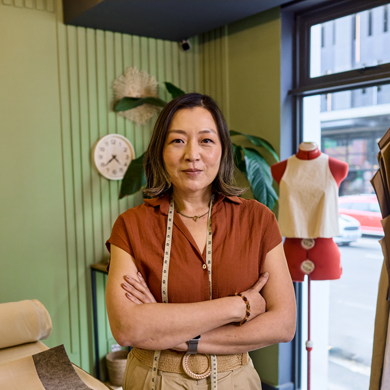 A single worker standing confidently with arms crossed in a tailoring workshop, surrounded by fabric rolls, a measuring tape around the neck, and clothing racks with garments